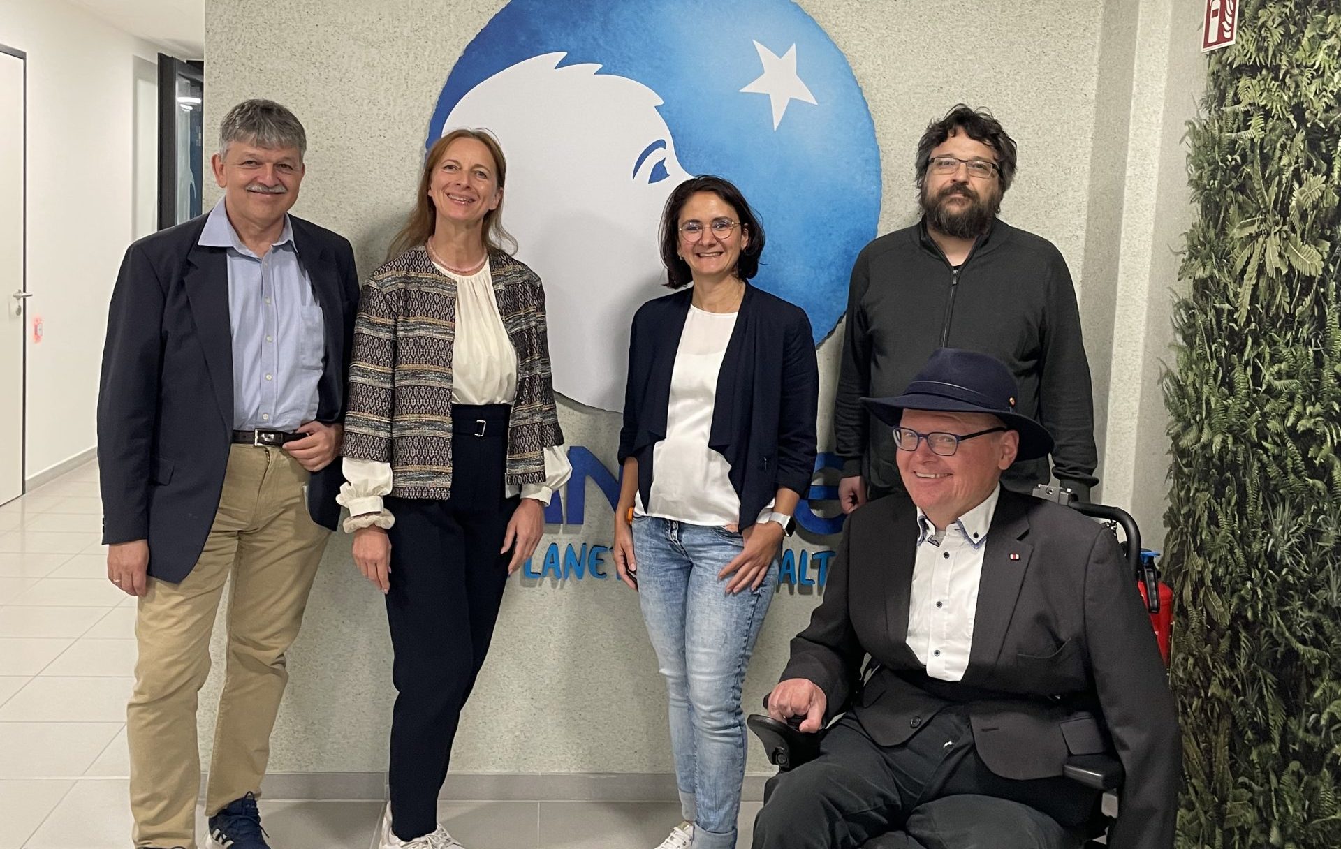 Bildbeschreibung: Die Workshopteilnehmenden Dietmar Stippler, Silke Kadach, Phyllis Ros, Michael Gubo und Karl-Heinz Miederer (v.l.n.r.) stehen vor der Wand mit blauem Danone Logo „One Planet. One Health.“ und blicken freundlich in die Kamera.